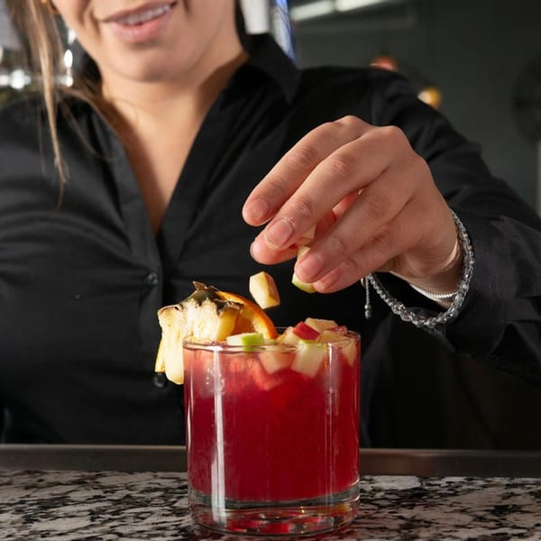 A bartender garnishes a red cocktail with diced fruit and an orange slice at Alma Kitchen.