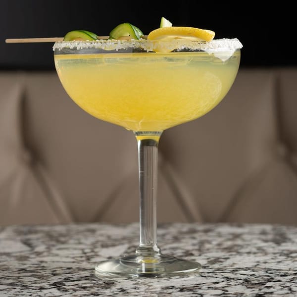 A vibrant yellow Alma Kitchen cocktail, garnished with cucumber and lemon, on a speckled countertop.