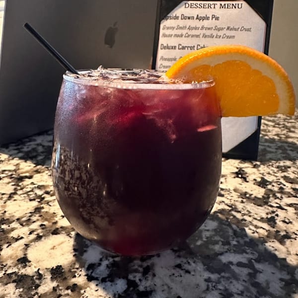 A dark red drink with ice, a black straw, and an orange slice on a speckled counter.