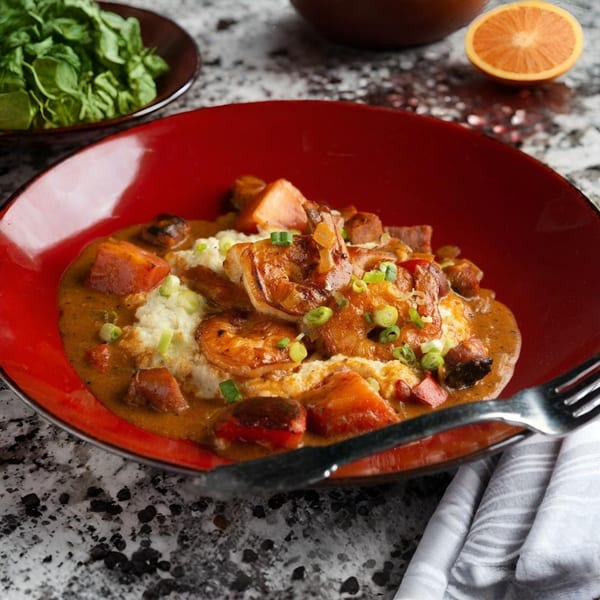 Shrimp and grits with sausage and sweet potatoes in a red bowl, with a side salad and orange.