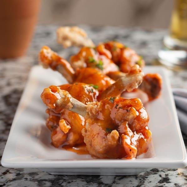 Alma Kitchen's chicken lollipops, coated in a glistening orange sauce and garnished with herbs, on a white plate.